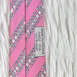 May include: A pink necktie featuring a repeating diagonal pattern of grey and white interlocking chain links. A rectangular label is stitched to the back, displaying the text: MADE IN FRANCE, 5402 OA, 100% SOIE. The fabric has a smooth, woven texture.