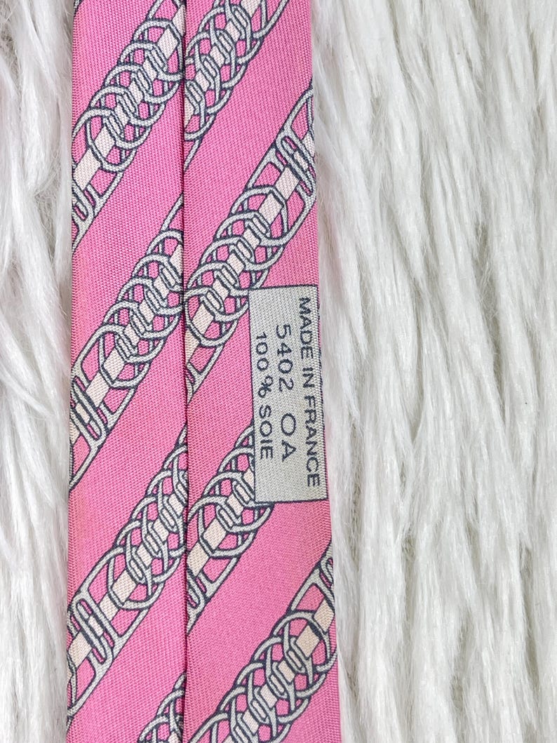 May include: A pink necktie featuring a repeating diagonal pattern of grey and white interlocking chain links. A rectangular label is stitched to the back, displaying the text: MADE IN FRANCE, 5402 OA, 100% SOIE. The fabric has a smooth, woven texture.
