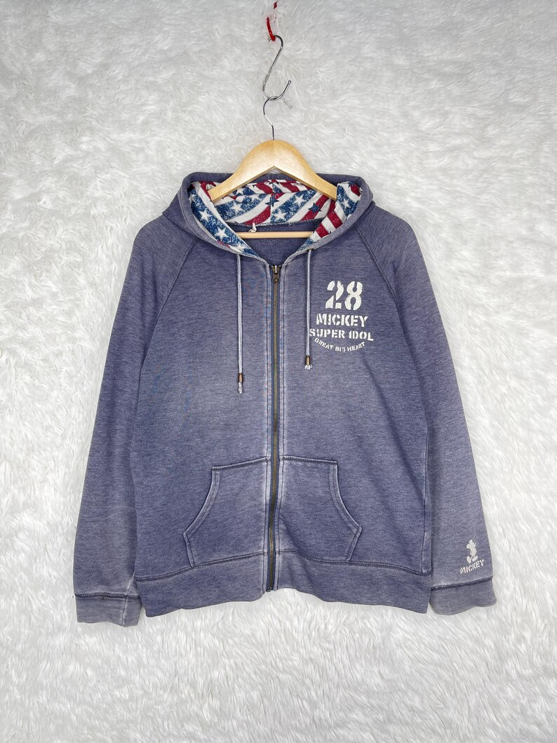 Może przedstawiać: Niebieska bluza z kapturem zapinana na zamek błyskawiczny, z przednią kieszenią i kapturem wyłożonym czerwono-biało-niebieskim wzorem. Bluza ma napis "28 MICKEY SUPER IDOL GREAT BIT HEART" na piersi i małą grafikę Myszki Miki.