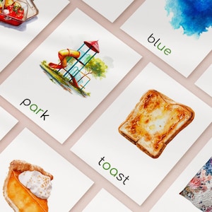 May include: A collection of watercolor flashcards featuring illustrations and words. Cards include a playground, toast, and the word "park". The cards are arranged on a light pink surface.
