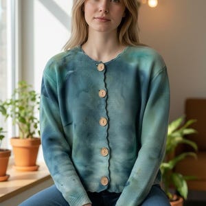 May include: A blue and green tie-dye cardigan with a round neckline and wooden buttons. The long-sleeved sweater is paired with blue jeans. The cardigan is a comfortable, casual style.