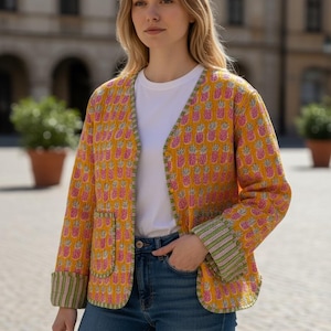 May include: A yellow jacket with a pink pineapple pattern. The jacket has a V-neck, two pockets, and green and white striped cuffs. The person is wearing a white t-shirt and blue jeans.