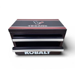 May include: A black and red toolbox with two drawers and a top featuring the Houston Texans logo and the word "TEXANS". The word "KOBALT" is printed in white on the front of the toolbox.