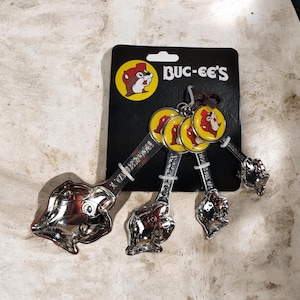 Könnte beinhalten: Ein Satz Buc-ee's Messlöffel. Die Löffel sind silberfarben mit einem Biberkopf-Design. Das Set enthält 1 Esslöffel, 1 Teelöffel, 1/2 Teelöffel und 1/4 Teelöffel. Die Löffel sind an einer schwarzen Karte mit dem Buc-ee's-Logo befestigt.