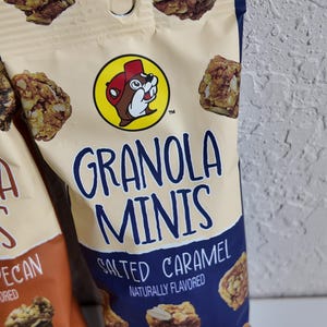 May include: A close-up of a bag of granola minis with the text "GRANOLA MINIS SALTED CARAMEL NATURALLY FLAVORED". The bag has a beige top and a navy blue bottom. The bag features a cartoon beaver logo.
