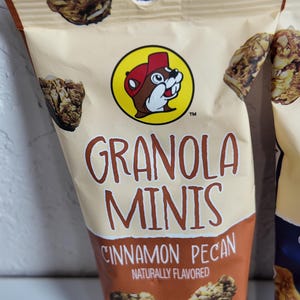 May include: A beige bag of granola minis with the text "GRANOLA MINIS CINNAMON PECAN NATURALLY FLAVORED". The bag features a cartoon beaver wearing a red hat. Granola pieces are visible on the bag.