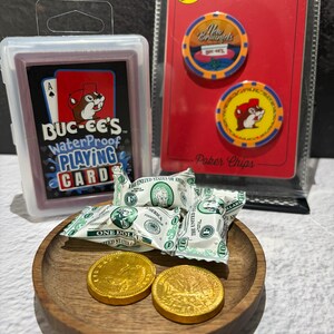 May include: A selection of Buc-ee's branded items. The image shows a deck of waterproof playing cards, two poker chips, wrapped sweets, and two gold-coloured coins, presented on a wooden tray.