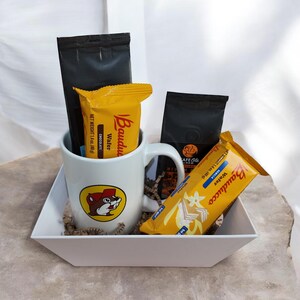 May include: A white mug with a cartoon beaver logo, containing a chocolate wafer, a vanilla wafer, and two black coffee packets, displayed in a white square tray.