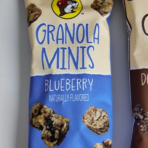 May include: A package of blueberry granola minis with the text "GRANOLA MINIS BLUEBERRY NATURALLY FLAVORED". The package has a blue and tan color scheme and features a cartoon beaver logo. The net weight is 2 oz (56g).