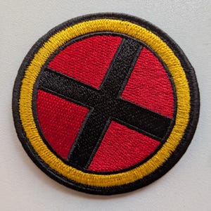 Può includere: Una toppa ricamata circolare con uno sfondo rosso vivace e una croce nera audace centrata. Il design è racchiuso da un bordo circolare spesso di colore giallo brillante, ulteriormente delineato da un sottile bordo nero cucito.