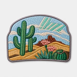 Può includere: Una toppa ricamata raffigurante un paesaggio desertico con un alto cactus saguaro verde, dune di sabbia dorata, una formazione rocciosa marrone e un piccolo cactus rotondo con fiori rosa sotto un cielo azzurro con nuvole e raggi verdi.
