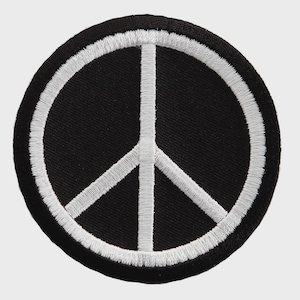 Puede incluir: Un parche bordado circular que presenta un símbolo de la paz blanco centrado sobre un fondo negro sólido. El diseño está encerrado por un grueso borde cosido en blanco, creando un elemento gráfico de alto contraste con acabado de hilo.