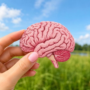 May include: An embroidered patch shaped like a human brain, featuring detailed stitching in shades of light pink and darker magenta. The design highlights the anatomical folds and structures of the brain, including the cerebellum in a darker pink.