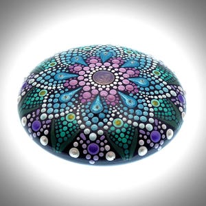 May include: A hand-painted, oval-shaped stone featuring a mandala design. The stone is primarily black, adorned with a symmetrical pattern of colorful dots in shades of blue, teal, purple, pink, and white. The design is centered with a metallic gold accent.
