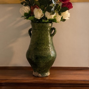 Op de afbeelding: Een donkergroene keramieken vaas met twee kleine handvatten aan de zijkant, voorzien van een glanzend, gevlekt glazuur. De vaas is gevuld met een arrangement van kunstbloemen, waaronder donkerrode rozen en witte bloesems met groene bladeren.