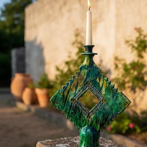 Könnte beinhalten: Ein grüner und goldener Kerzenhalter aus Keramik mit rautenförmigem Design und einer brennenden weißen Kerze. Der Kerzenhalter hat eine strukturierte Oberfläche und einen dekorativen Sockel. Die Kerze ist etwa 20 cm hoch.