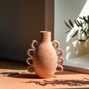 May include: A terracotta-colored vase with a unique design. The vase has a rounded body and a narrow neck. It features six loop handles evenly spaced around the body. The vase is displayed on a terracotta-colored surface, with shadows cast by sunlight.