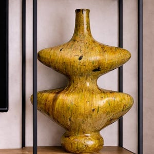 May include: A large, decorative vase with a distinctive layered design. The vase is mainly yellow with dark brown and black accents, giving it a textured appearance. It sits on a wooden shelf within a black metal frame.