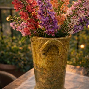 May include: A tall, olive-green ceramic vase with two handles holds a vibrant bouquet of colorful flowers. The flowers display shades of red, pink, purple, and orange. The vase has a textured surface and sits on a wooden table.