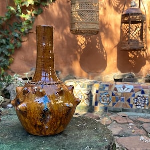 May include: A brown ceramic vase with a textured finish and a distinctive, rounded design. The vase features a slender neck and is decorated with small, spherical bumps. It rests on a moss-covered stone, with a blurred background of hanging lanterns.