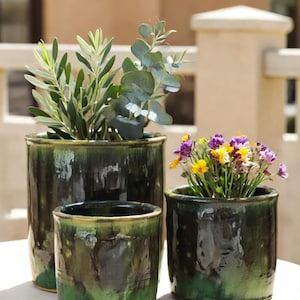 May include: Three glazed ceramic planters in varying sizes, each with a dark green and yellow color scheme. The planters hold greenery and colorful flowers, showcasing a rustic aesthetic. The largest planter contains olive branches.