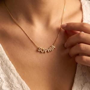 Könnte beinhalten: Eine goldfarbene Halskette mit dem Namen "SOPHIA" in einzelnen Großbuchstaben. Die Halskette wird um den Hals getragen und liegt auf einem weißen Spitzentop. Die Buchstaben sind durch eine filigrane Kette verbunden.