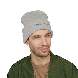 May include: A gray beanie with the text "Pacific Northwest Bell" in blue. The beanie is made of a soft material and is worn by a person wearing a green bomber jacket.
