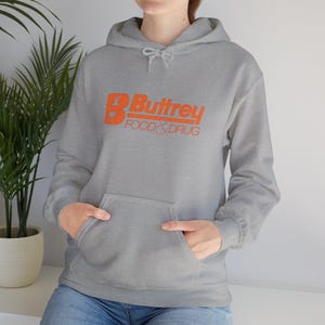 Puede incluir: Una sudadera con capucha gris con un bolsillo delantero y texto naranja que dice "B Buttrey FOOD & DRUG". La sudadera tiene una capucha con cordón y puños acanalados. El modelo lleva vaqueros azules.