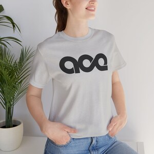 Może przedstawiać: Jasnoszara koszulka z napisem "ace" w czarnym, stylizowanym kroju pisma. Koszulka z krótkim rękawem jest wykonana z miękkiego materiału. Osoba ma na sobie niebieskie dżinsy.