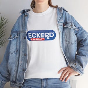 Puede incluir: Camiseta blanca con el logo de Eckerd Pharmacy. El logo es un óvalo azul con la palabra "ECKERD" en blanco, un símbolo "Rx" rojo y la palabra "PHARMACY" en rojo. La persona lleva una chaqueta vaquera.