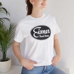 May include: White t-shirt featuring the Eisner Food Stores logo in a black oval. The logo includes the word "Eisner" in a stylized script above "Food Stores". The shirt has short sleeves and a crew neck.