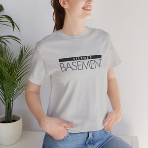 Peut inclure: Un t-shirt gris clair à manches courtes avec le texte noir Filene's Basement imprimé sur la poitrine. Le motif comprend une barre horizontale noire solide au-dessus du mot Filene's et le mot Basement dans une police sans empattement.