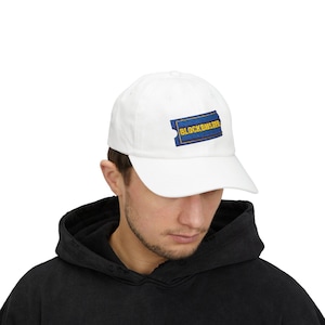 May include: White baseball cap with a Blockbuster logo patch. The patch is a blue rectangle with a yellow border and the word "BLOCKBUSTER" in yellow. The cap is made of a soft material and has a curved peak.