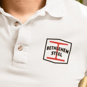 以下が含まれることがあります： クラシックな襟と2つの茶色のボタンが付いた、明るいグレーのポロシャツ。シャツには、大胆で様式化されたフォントで「BETHLEHEM STEEL」と書かれた黒と赤のロゴが付いています。シャツはテクスチャード加工された生地で作られています。