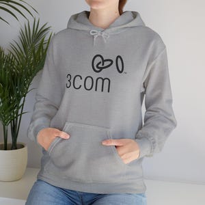Puede incluir: Una sudadera con capucha gris claro con un bolsillo delantero y una capucha con cordón. La sudadera con capucha presenta el texto negro "3COM" y un logotipo estilizado encima del texto. La persona lleva vaqueros azules.
