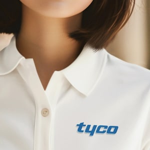 以下が含まれることがあります： 襟とボタン付きの前立てが付いた白いポロシャツ。シャツの前面には青い文字で「tyco」と書かれています。モデルは短い茶色の髪をしており、シャツを着ています。