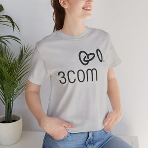Puede incluir: Una camiseta gris claro con el texto negro "3COM" y un gráfico estilizado. La camiseta de manga corta parece estar hecha de un material suave. El diseño está centrado en la parte delantera de la camiseta.