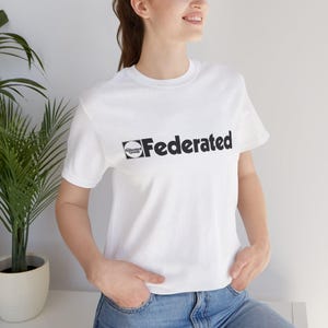 Puede incluir: Una camiseta blanca con la palabra "Federated" impresa en negro, junto con un pequeño logotipo circular. La camiseta tiene mangas cortas y cuello redondo. La persona lleva vaqueros azules.