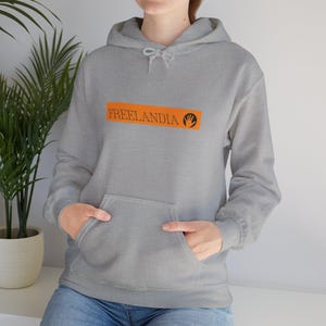 May include: A gray hooded sweatshirt with a front pocket and drawstring. The sweatshirt features the word "FREELANDIA" in orange block letters with a black hand graphic. The model is wearing blue jeans.