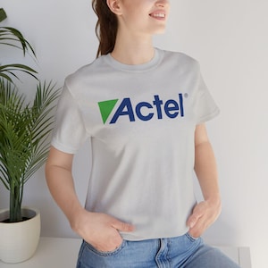 Könnte beinhalten: Ein hellgraues T-Shirt mit dem Wort "Actel" in Blau und einem grünen Dreieck. Das kurzärmelige Shirt ist aus einem weichen Material. Die Person trägt eine blaue Jeans.