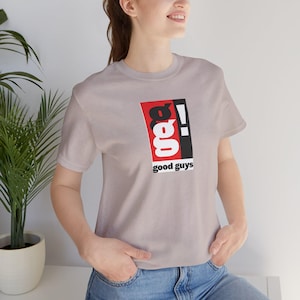 May include: A light grey short-sleeve t-shirt featuring a rectangular graphic on the chest. The graphic contains the letters g, g, and an exclamation point in black and white, set against a red and black background with the text good guys below.