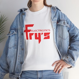 Puede incluir: Camiseta blanca con el texto rojo "Fry's Electronics" en una fuente audaz y estilizada. La camiseta se usa con una chaqueta vaquera azul claro. El look general es casual y cómodo.