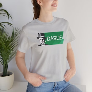 May include: A light gray t-shirt featuring a graphic of a man in a top hat and bow tie, with the word "DARLIE" in white letters on a green background. The shirt is made of a soft material.