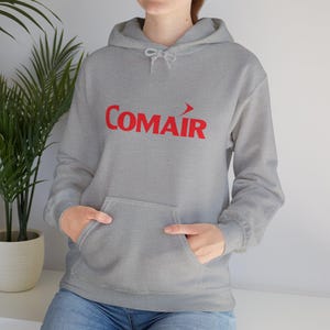 May include: A gray hooded sweatshirt with a front pocket and red text that reads "COMAIR". The sweatshirt has a drawstring hood and long sleeves. The person is wearing blue jeans.