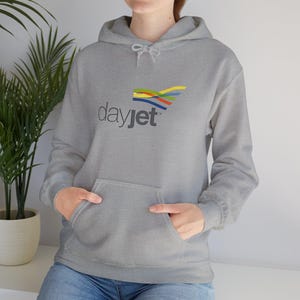 May include: A grey hooded sweatshirt featuring a front pocket and the word "dayjet" printed in dark grey. Above the text is a colourful abstract design. The sweatshirt is made of a soft material.