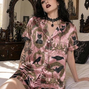Op de afbeelding: Roze satijnen pyjamaset met een herhalend patroon van ingelijste zwarte katten, kraaien en sierlijke gouden frames met rozen. De top met korte mouwen heeft een knoopsluiting en een borstzak. De shorts maken de set compleet.