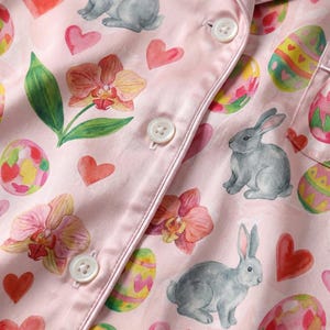 Puede incluir: Primer plano de tela de pijama rosa con tem&aacute;tica de Pascua. La tela presenta ilustraciones de acuarela de conejitos grises, huevos de Pascua decorados, corazones rojos y orqu&iacute;deas rosas. Botones blancos y ribete de sat&eacute;n visibles.