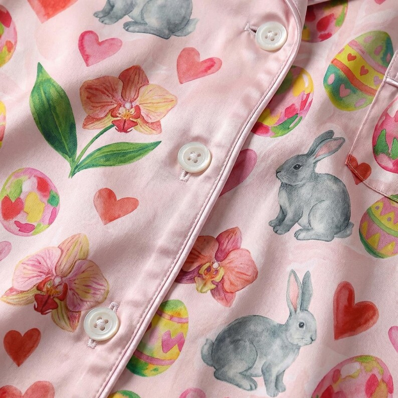 Puede incluir: Primer plano de tela de pijama rosa con tem&aacute;tica de Pascua. La tela presenta ilustraciones de acuarela de conejitos grises, huevos de Pascua decorados, corazones rojos y orqu&iacute;deas rosas. Botones blancos y ribete de sat&eacute;n visibles.