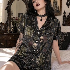 Op de afbeelding: Zwarte satijnen pyjamaset met een Medusa- en schedelprint. De top met korte mouwen heeft witte biezen en knopen. De shorts hebben een bijpassende print en witte rand. De set is ontworpen voor een gotische esthetiek.