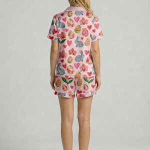 Puede incluir: Conjunto de pijama rosa con tem&aacute;tica de Pascua, con una camisa de manga corta y pantalones cortos a juego. El conjunto est&aacute; adornado con un patr&oacute;n repetido de huevos de Pascua, conejos grises, corazones rosas y flores. Los pijamas son de tela suave y ligera.